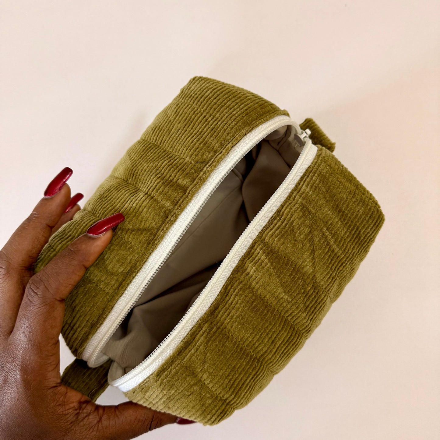 Green corduroy pouch held by a hand with red nail polish on a beige background
